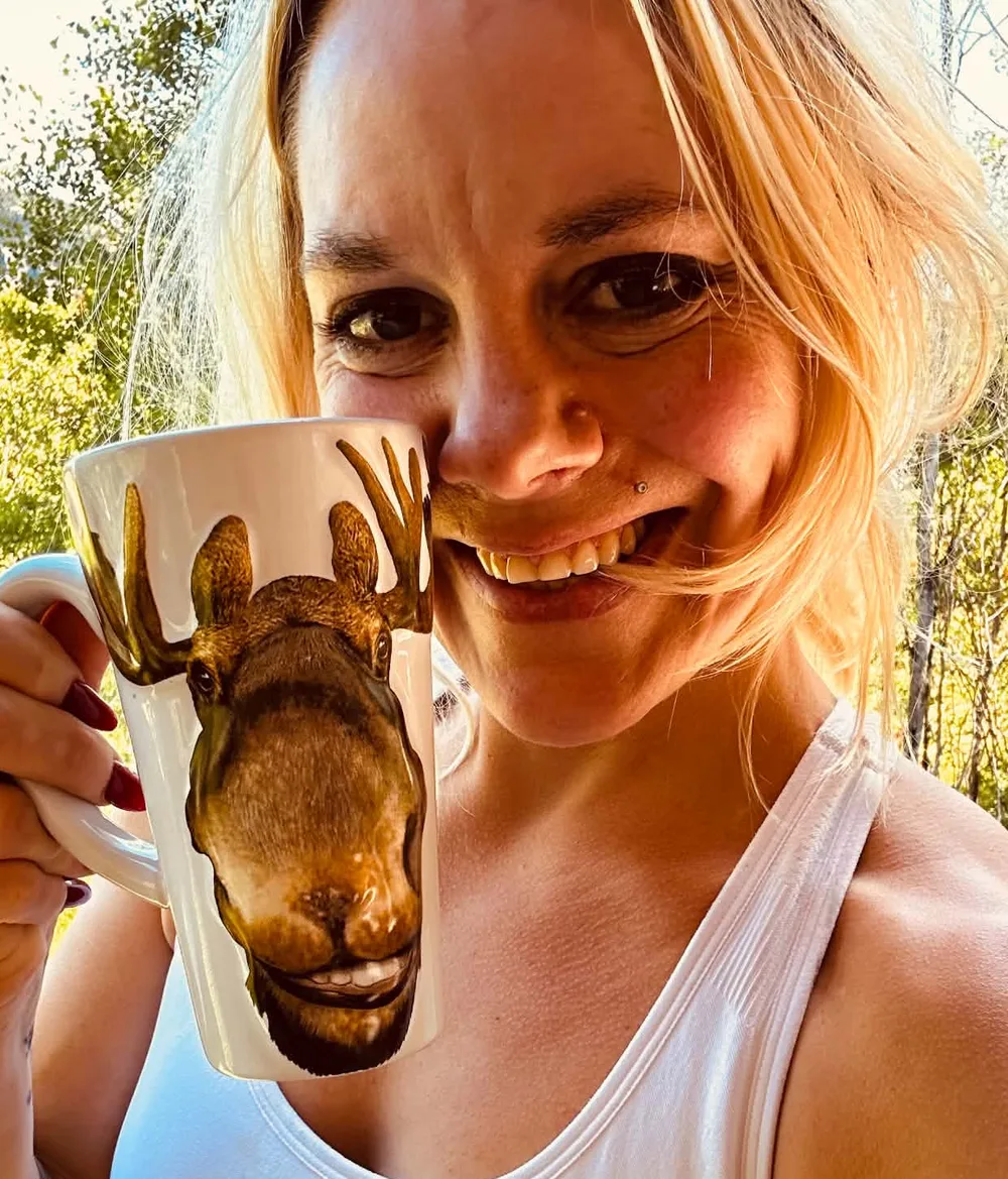 Jillian Mood smiling on a sunny deck, holding a tall mug with a moose on it